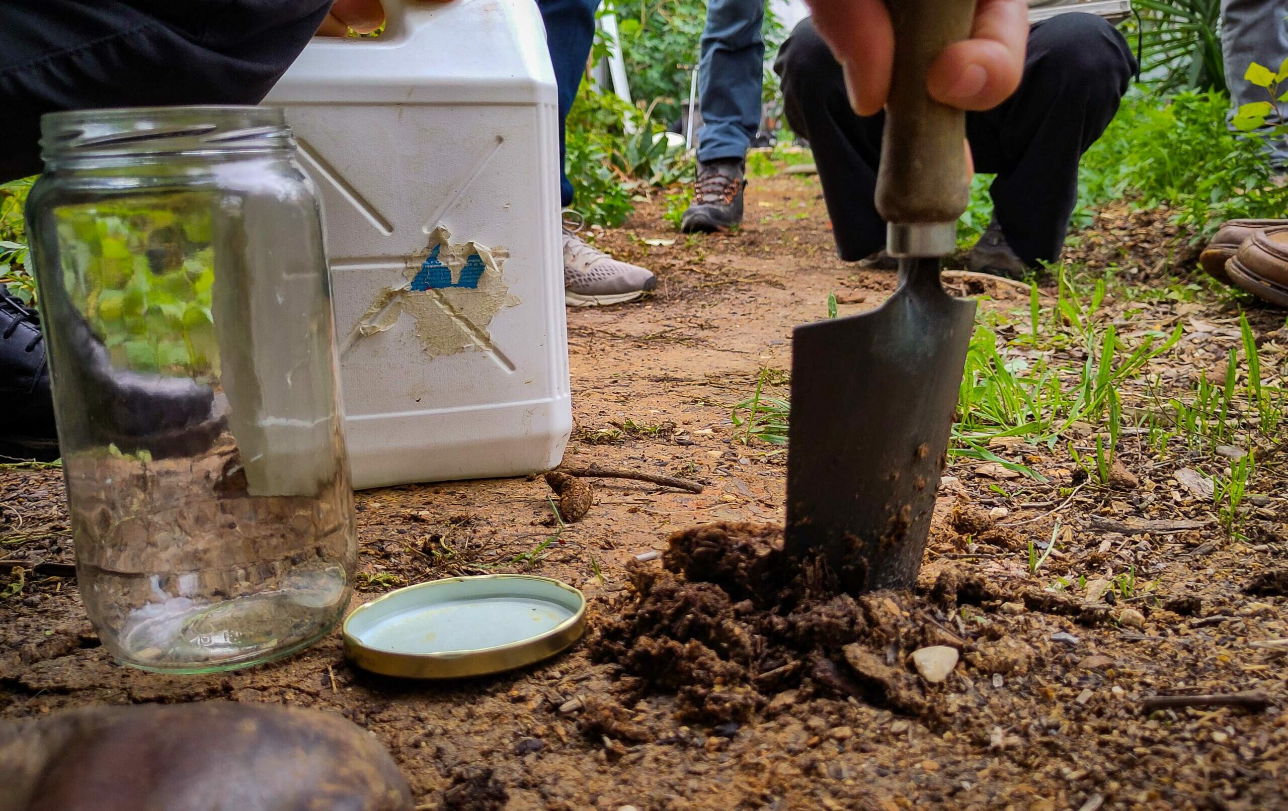 En gros plan un échantillonnage de sol à l'aide d'un truelle, s'u bocal et d'un bidon. En arrière plan, les jambes du public bénéficiaire de la formation qui rgarde l'échantillonage