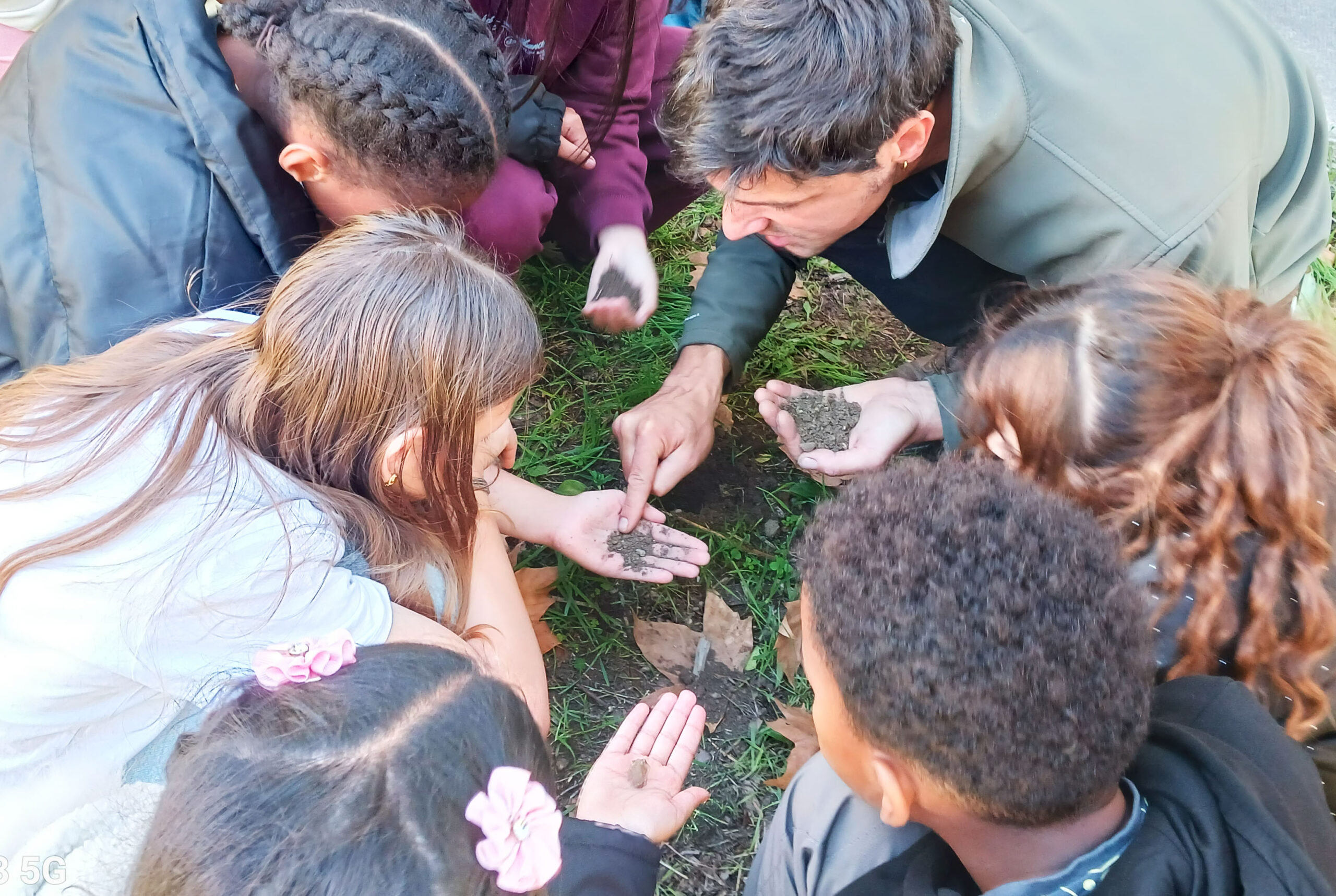 Des enfants rassembles en cercle autour d'une tranchee de sol le long d'un mur, avec un bidon d'eau et des bassines pour observer l'infiltration
