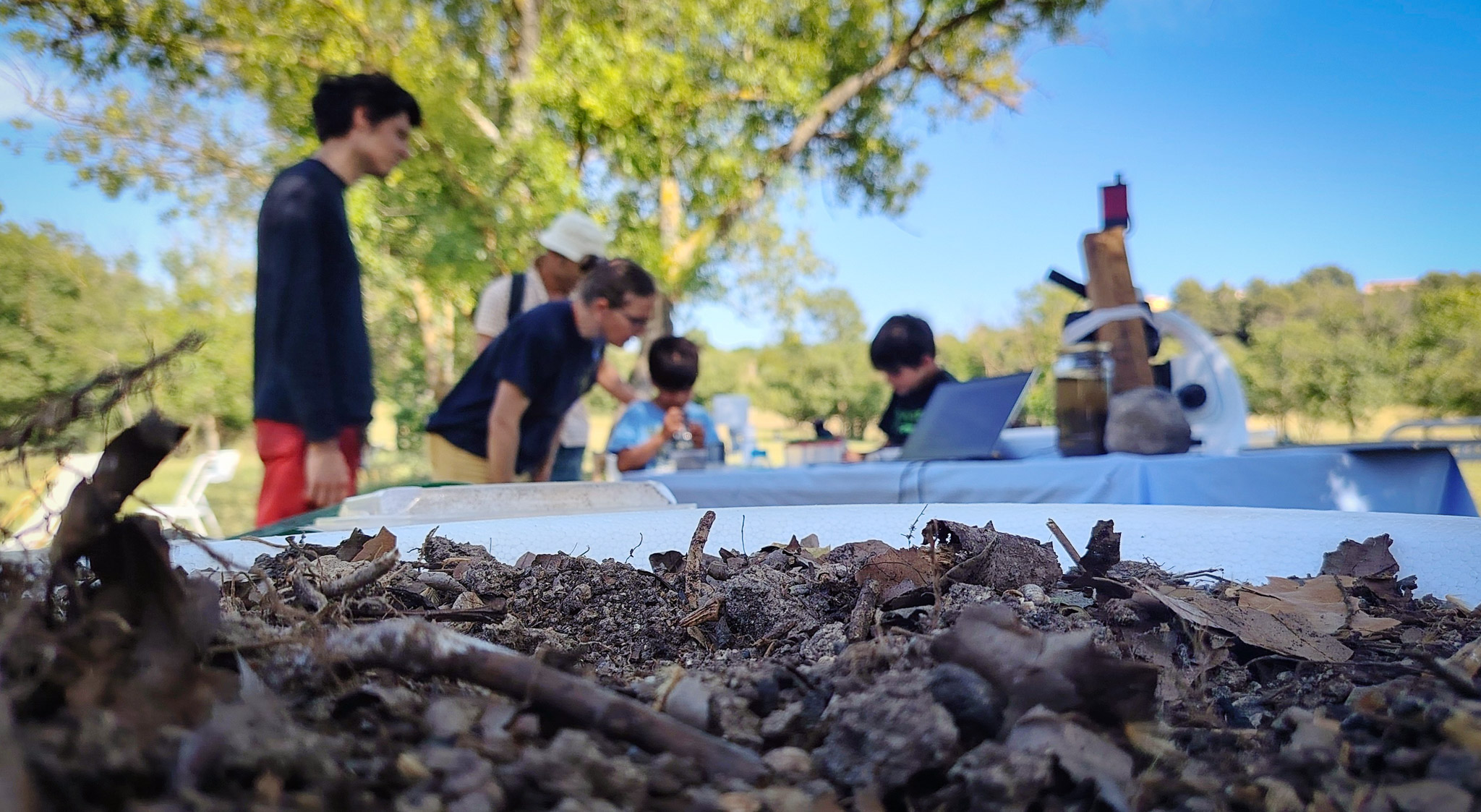 Au premier plan un échantillon de terre, en arrière plan un public familial qui observ la biodiversité avec des microscopes